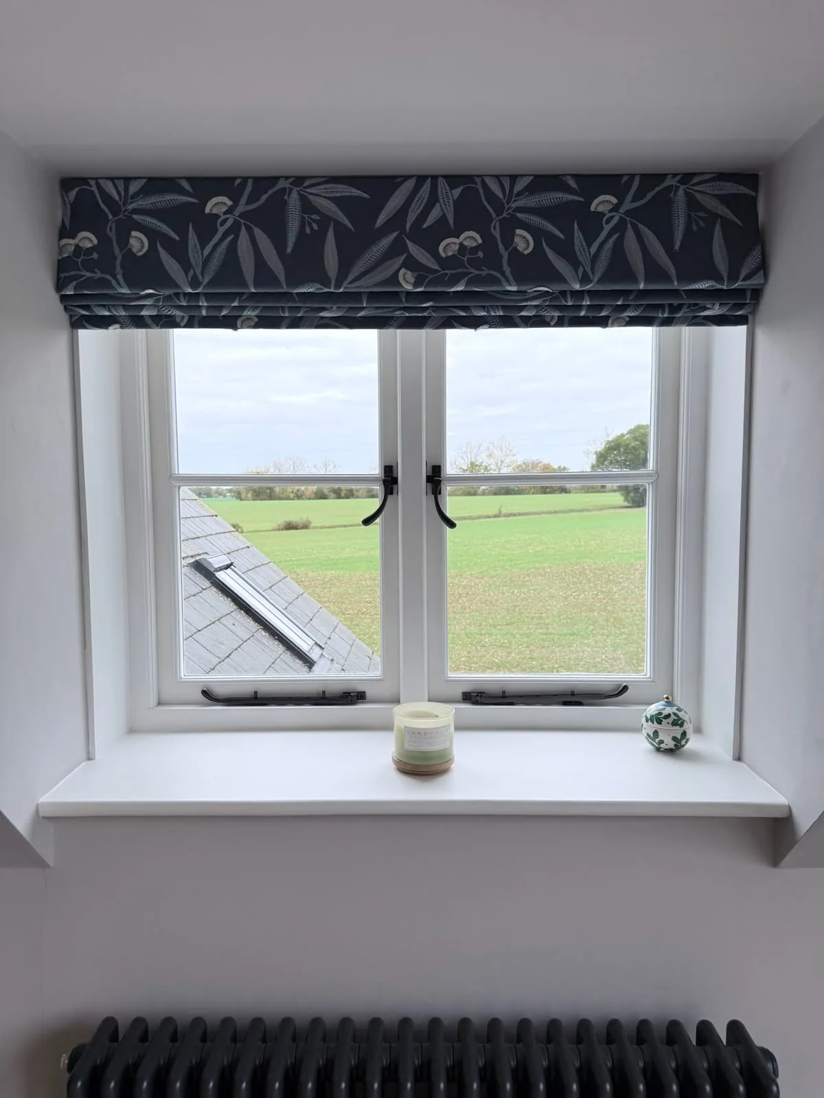 Charcoal botanical-print Roman blind raised in a deep white window reveal overlooking fields