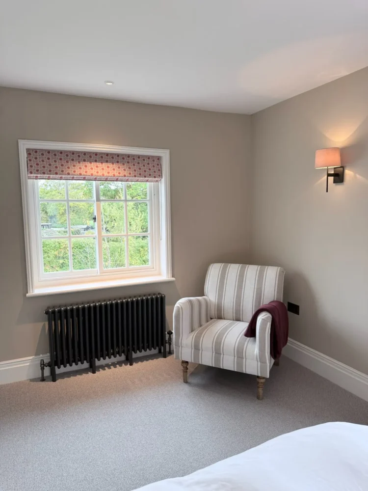 Bedroom corner with a patterned Roman blind at a sash window, a striped armchair and a cast-iron radiator