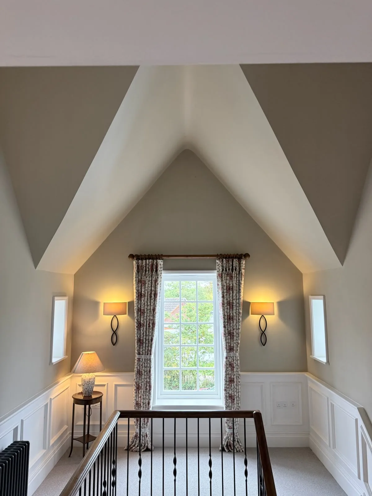 Gabled landing with floral curtains at a tall window between wall lights, above a staircase balustrade