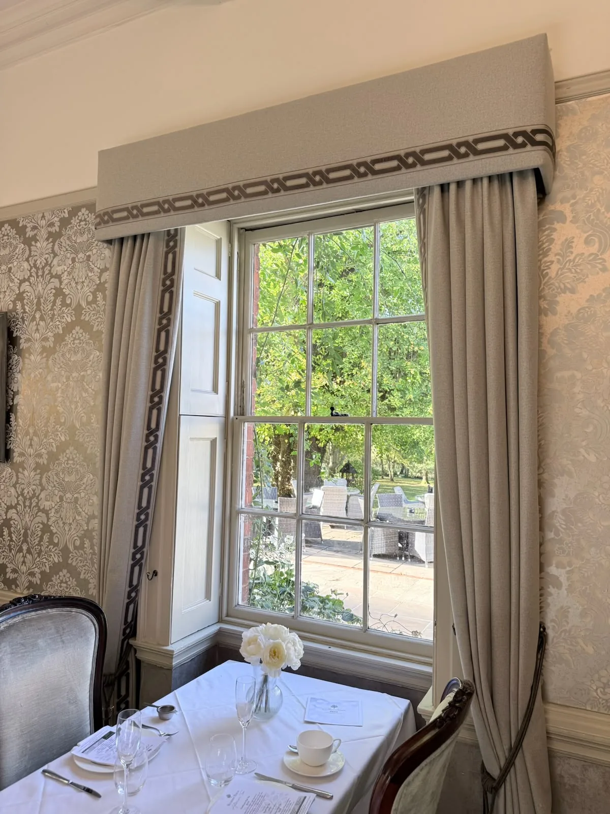 Trimmed upholstered pelmet and full-length curtains at a sash window, above a laid dining table