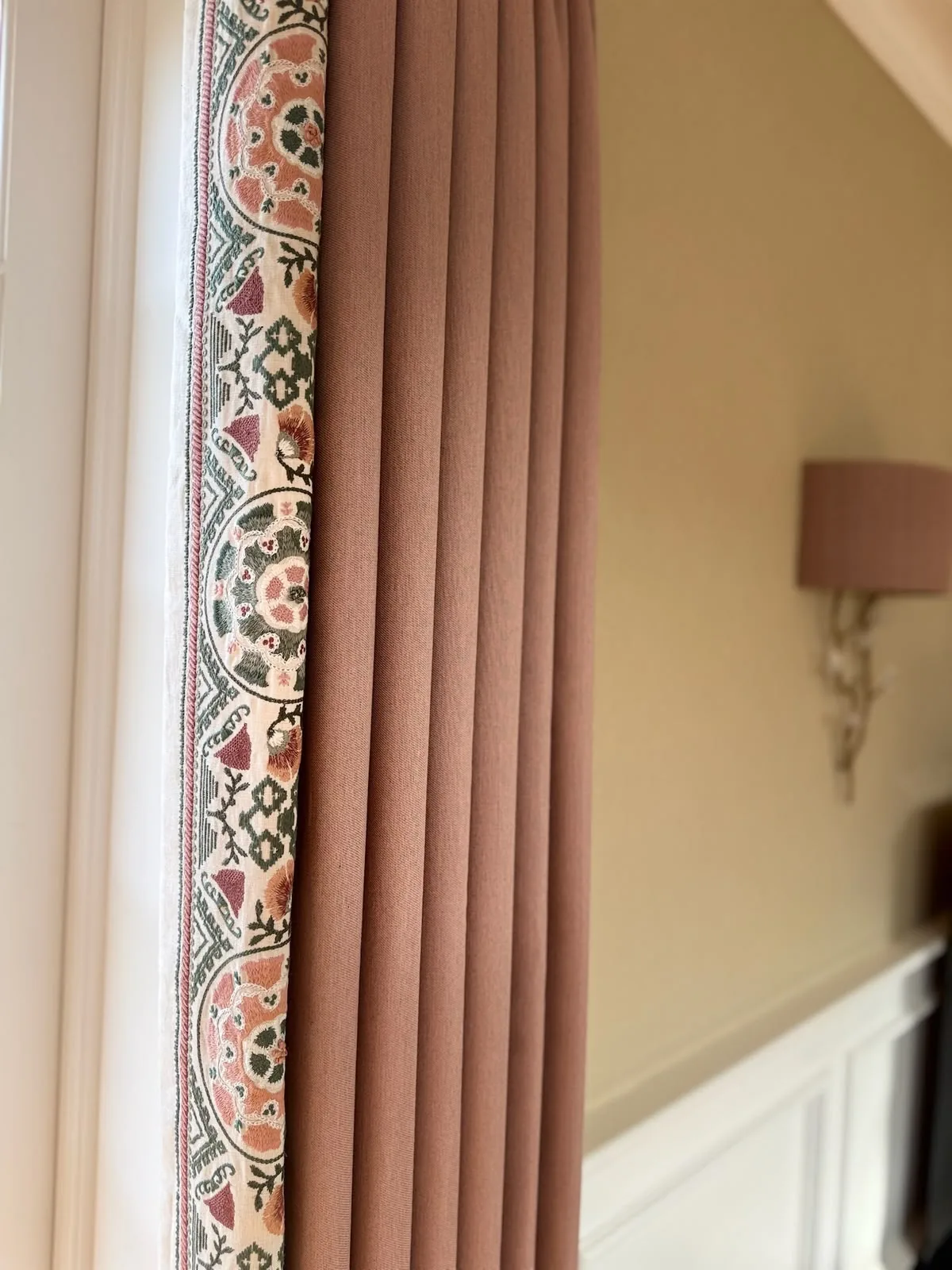 Dusky pink curtain with a printed floral border, hanging beside a window and a wall light