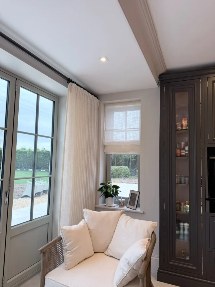 Cream curtains beside a door and window with a sheer Roman blind, a white armchair and a dark display cabinet