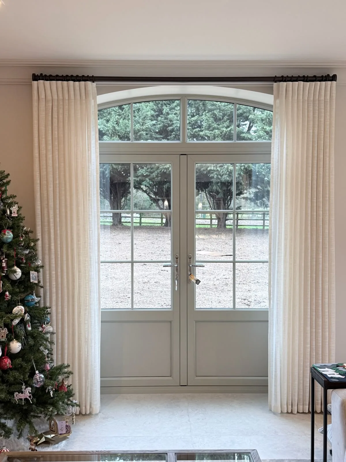 Cream curtains on a dark pole framing arched French doors, with a decorated Christmas tree to one side