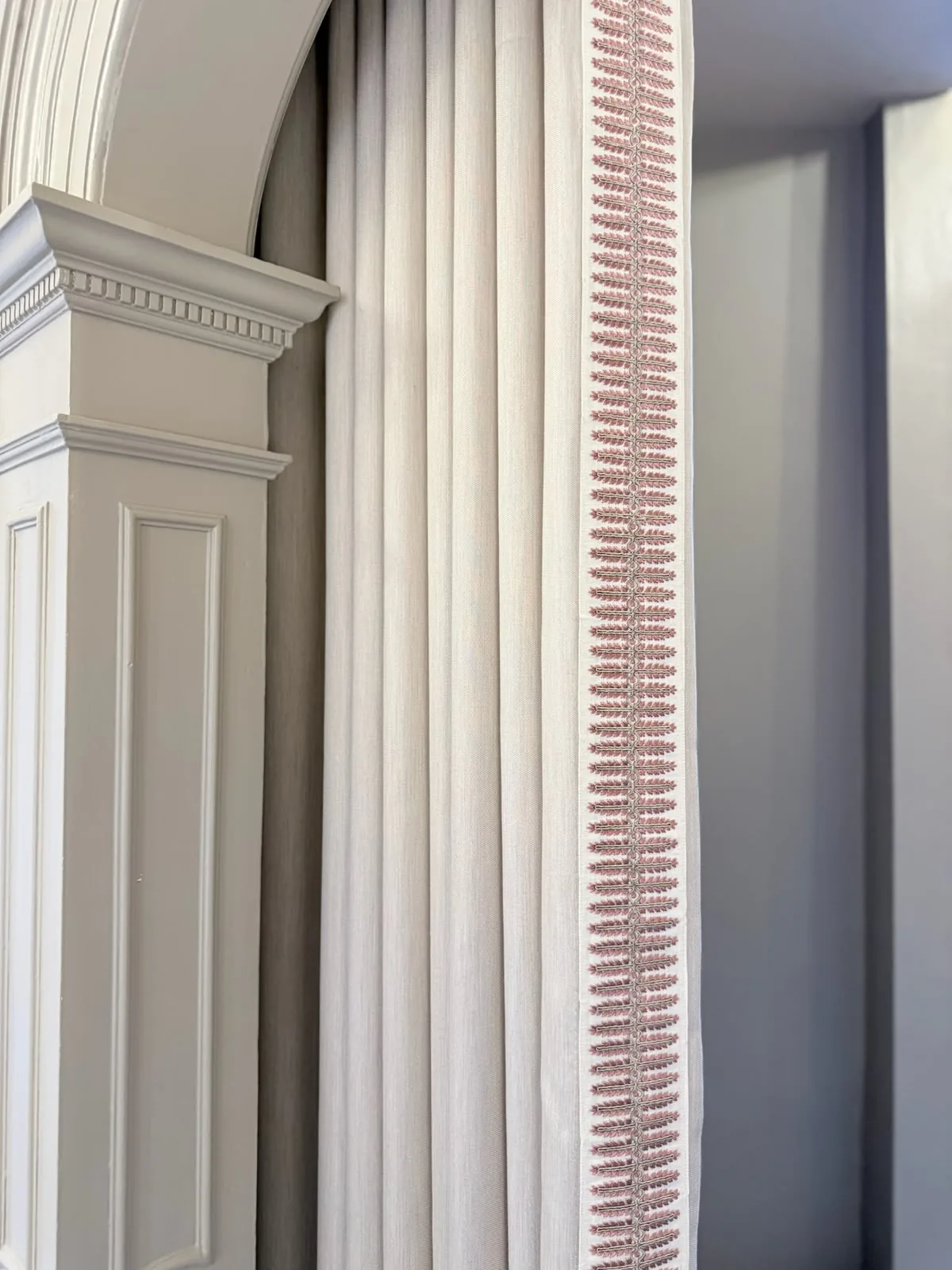 Ivory curtain with a pink leaf braid on the leading edge, hanging beside a fluted pilaster and arch