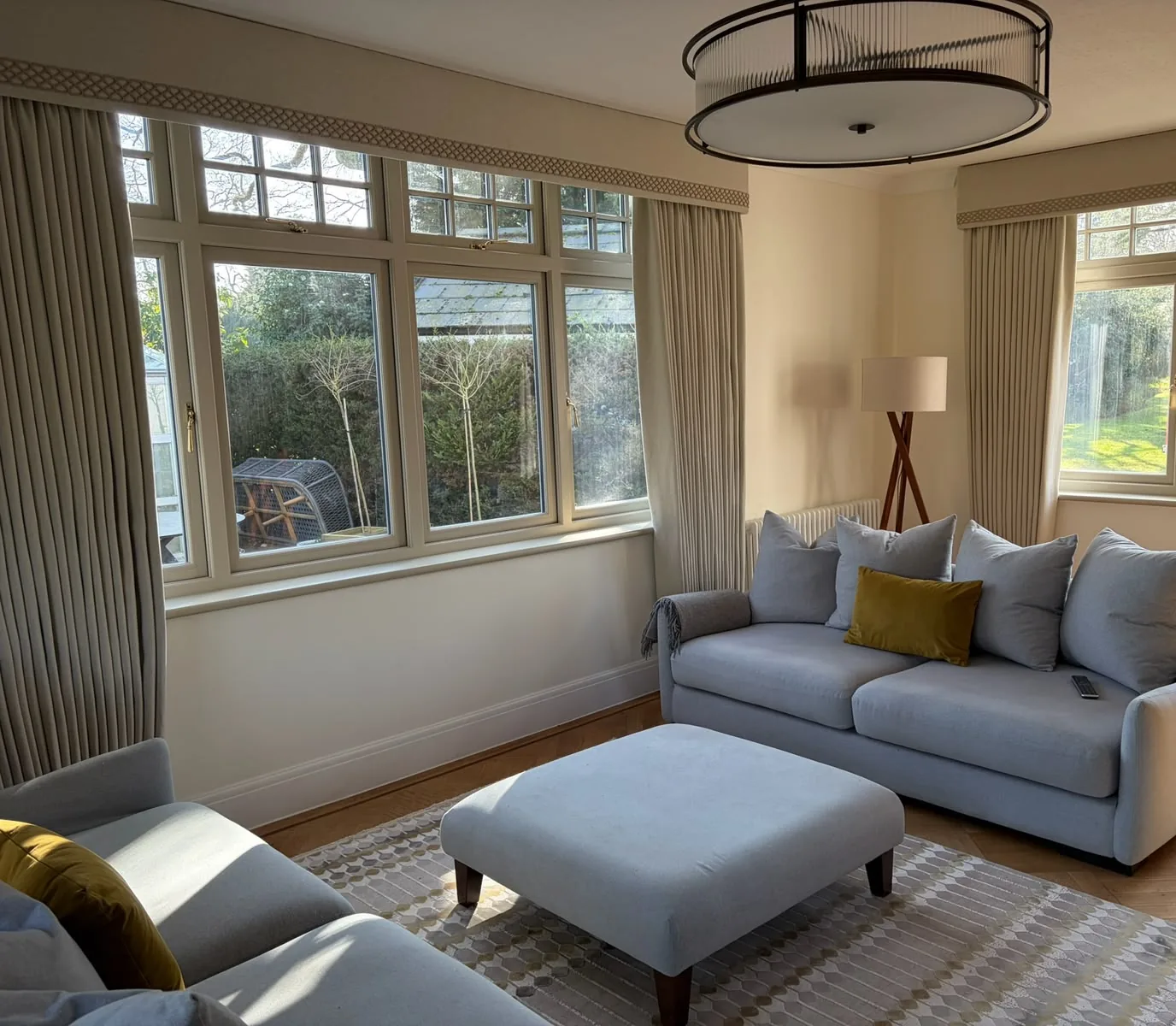 Sitting room with full-length neutral curtains under trimmed pelmets at two corner windows, a grey sofa and a matching ottoman