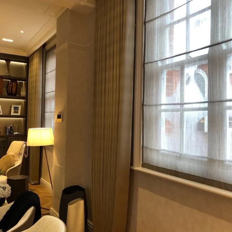 Ceiling-hung taupe curtains and sheer Roman blinds at tall windows, in a study with lit shelving and a floor lamp