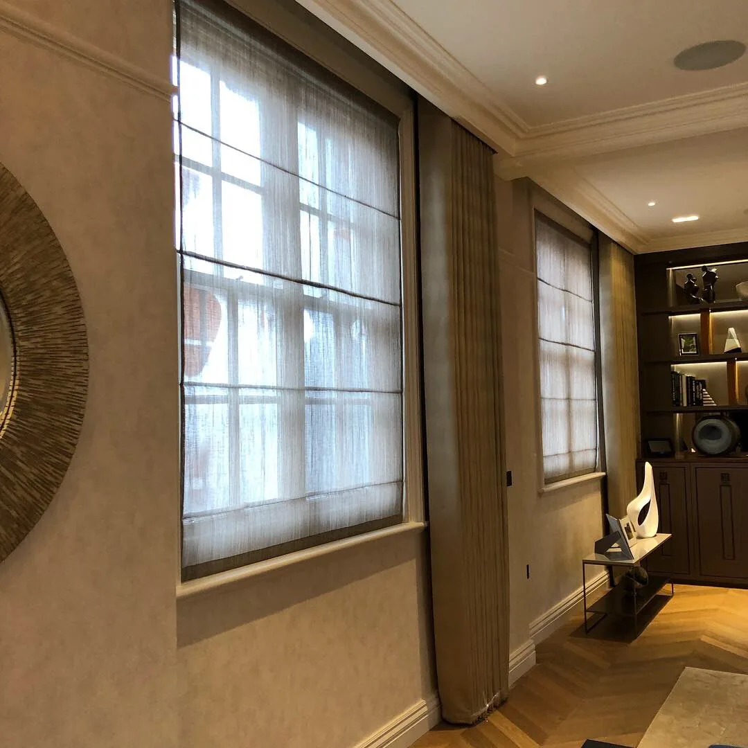 Sheer Roman blinds and ceiling-hung taupe curtains at a row of tall windows beside lit shelving