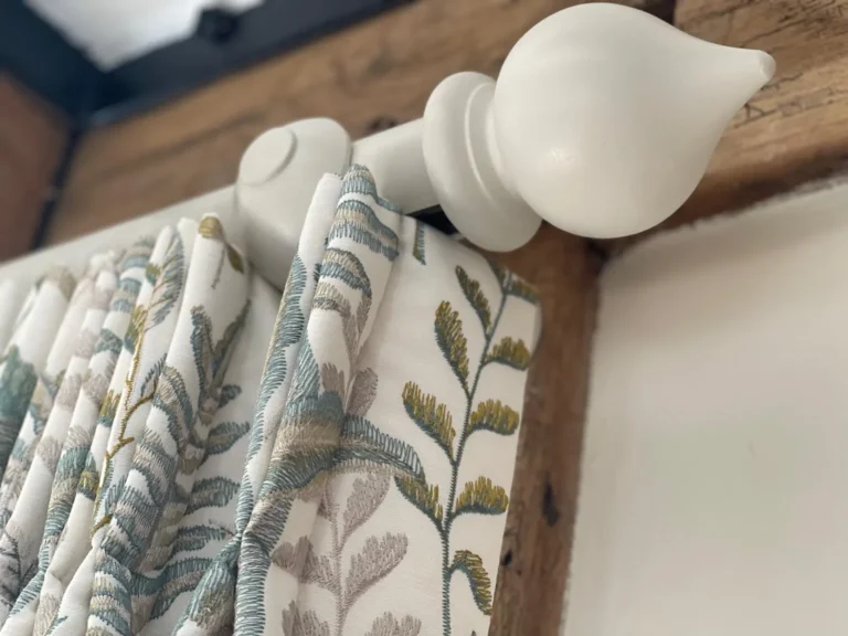 White wooden curtain pole with a turned finial, holding a leaf-print pinch-pleat curtain beside a beam