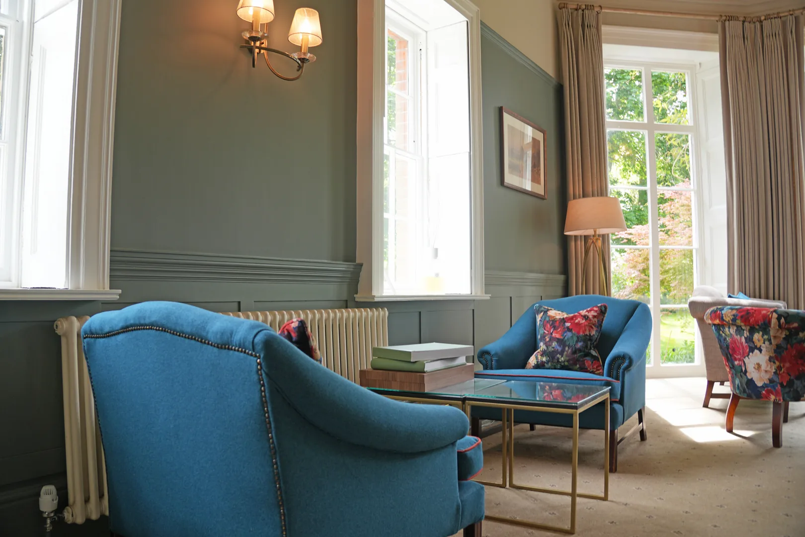 Panelled sitting room at Mulberry House with a blue upholstered sofa and armchair, floral velvet tub chairs and full-length curtains at tall sash windows