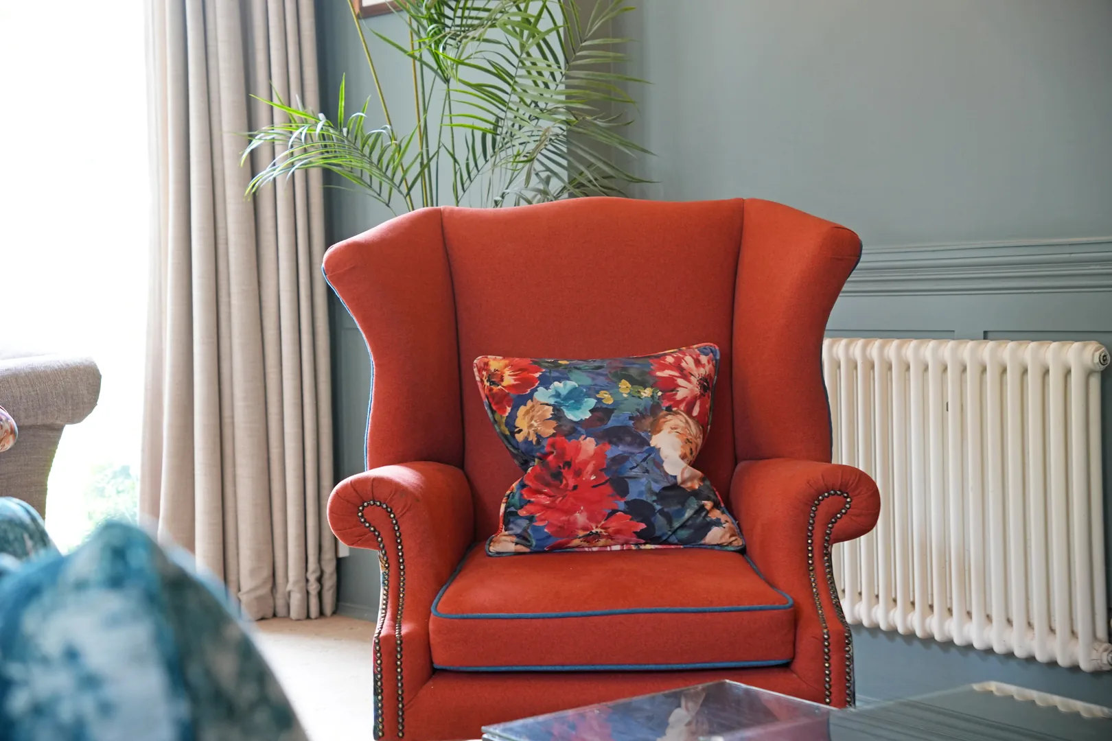 Rust velvet wingback armchair with stud detailing and a floral cushion, in the lounge at Mulberry House
