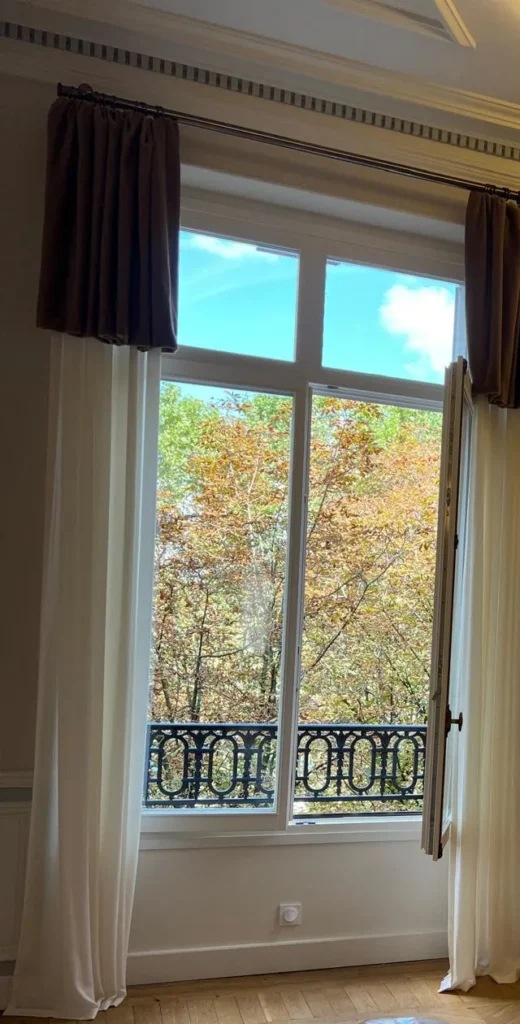 Tall French window dressed with sheer curtains and dark velvet top panels on a pole, opening onto a balcony and autumn trees