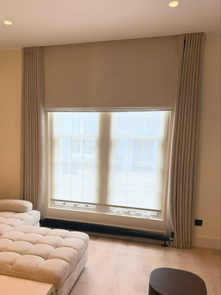 Neutral Roman blind lowered over a large window between floor-length curtains, beside a tufted sofa