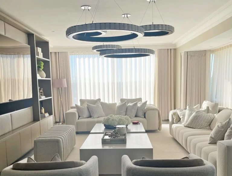 Large living room with sheer and dress curtains along a wall of windows, cream sofas and three ring pendant lights