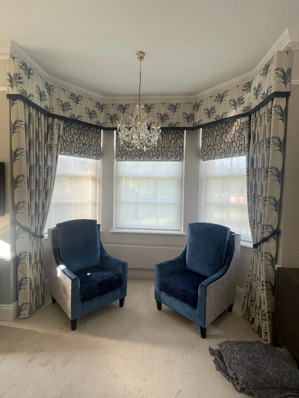 Bay window dressed with a shaped pelmet, fern-print curtains with tiebacks and Roman blinds, with two blue velvet armchairs