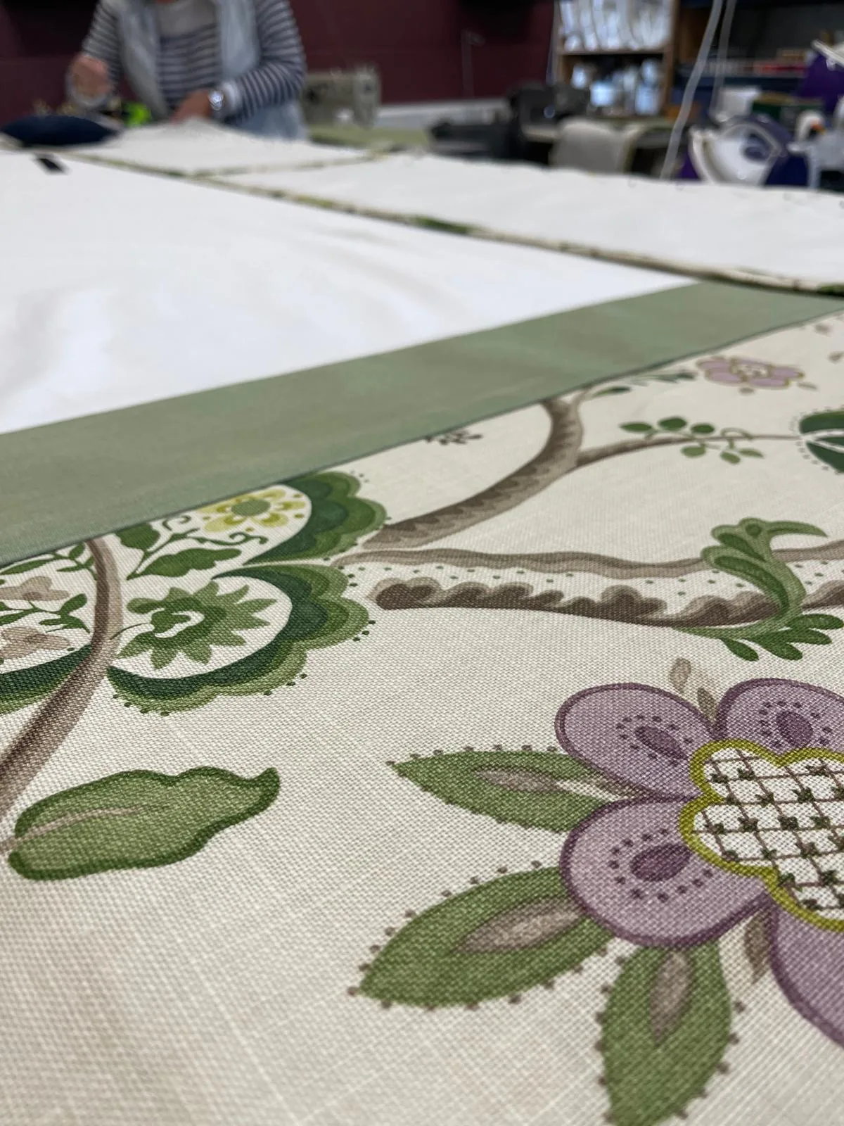 Floral crewel-style fabric with a plain green border being made up on the workroom table, with sewing machines behind