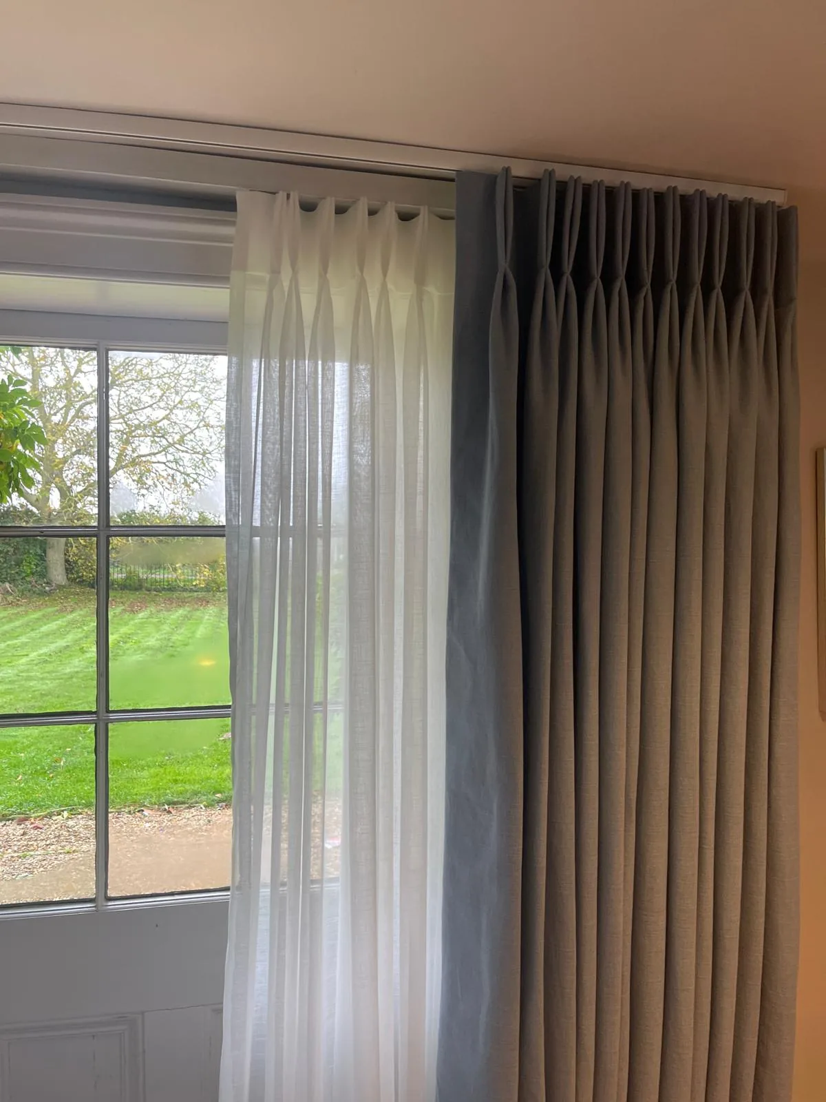 Blue-grey pinch-pleat curtain and a sheer voile on a double track at a window onto a striped lawn