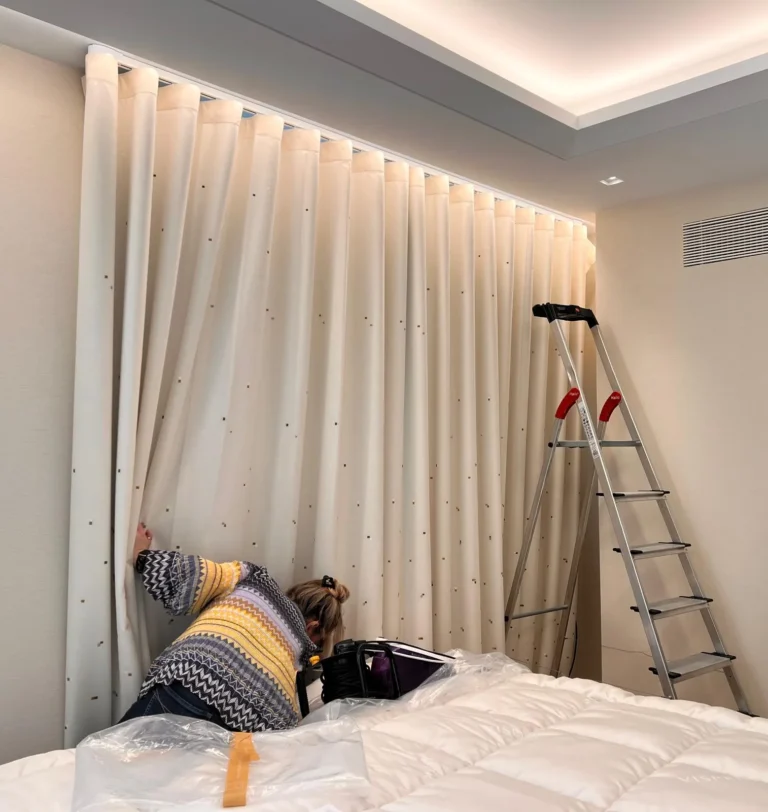 Sheer wave curtain being dressed on a recessed ceiling track, the fitter seen from behind beside a protected bed