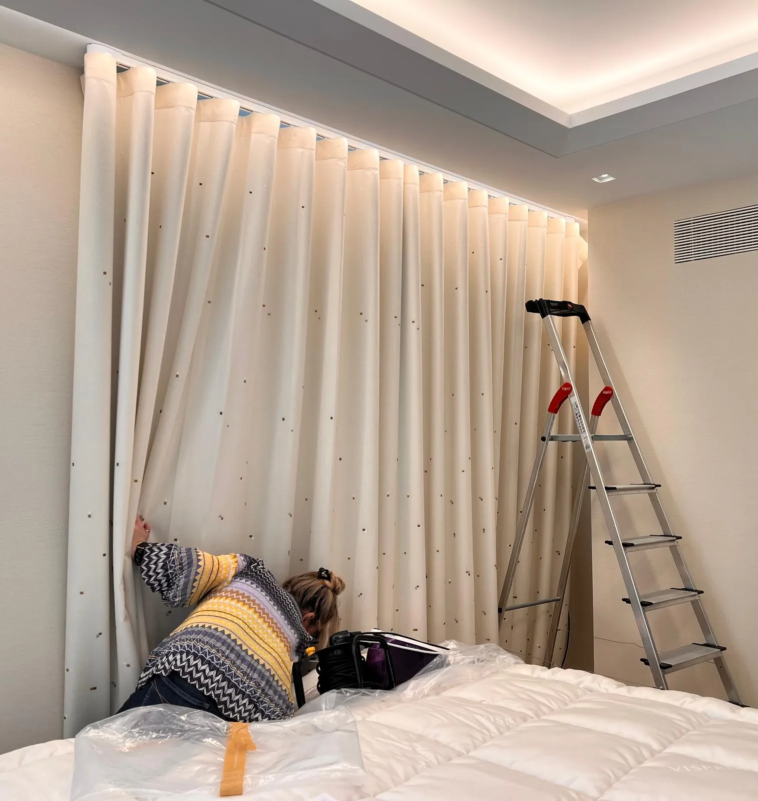 Sheer wave curtain being dressed on a recessed ceiling track, the fitter seen from behind beside a protected bed