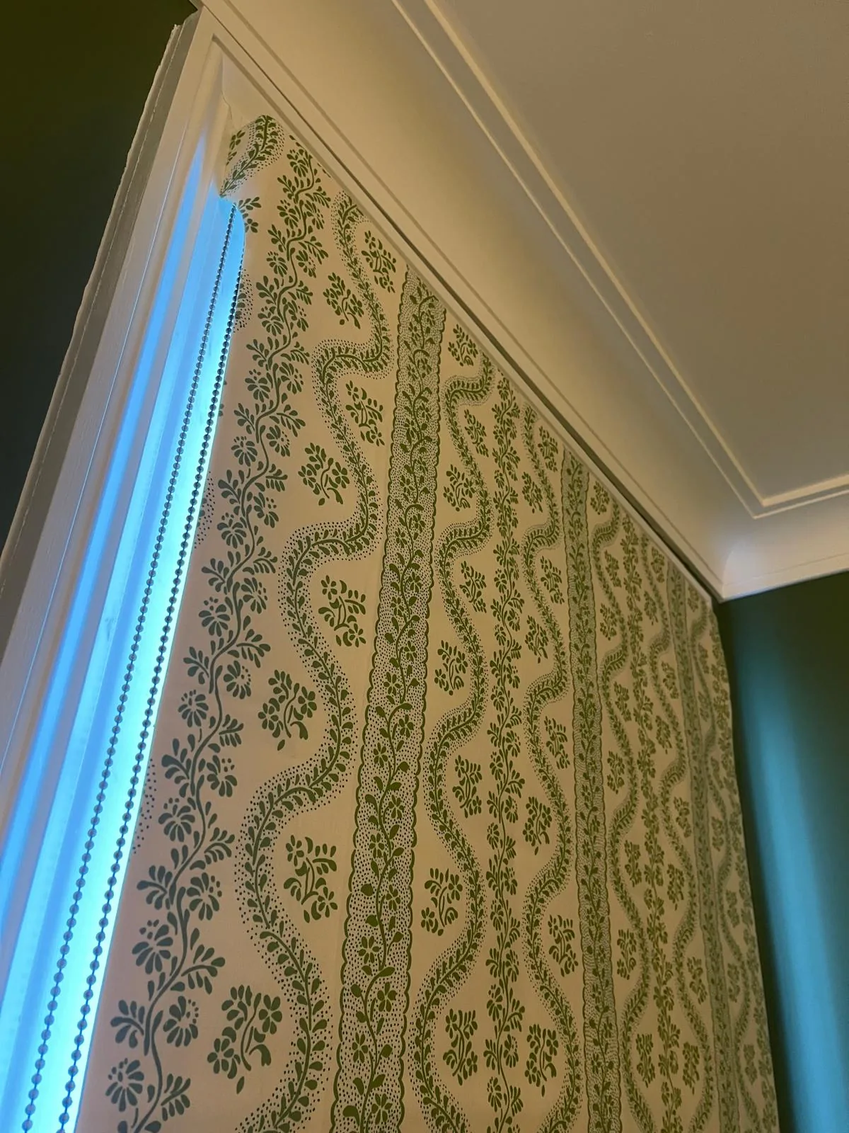 Green and white trellis-print Roman blind, seen from the side against a dark green wall