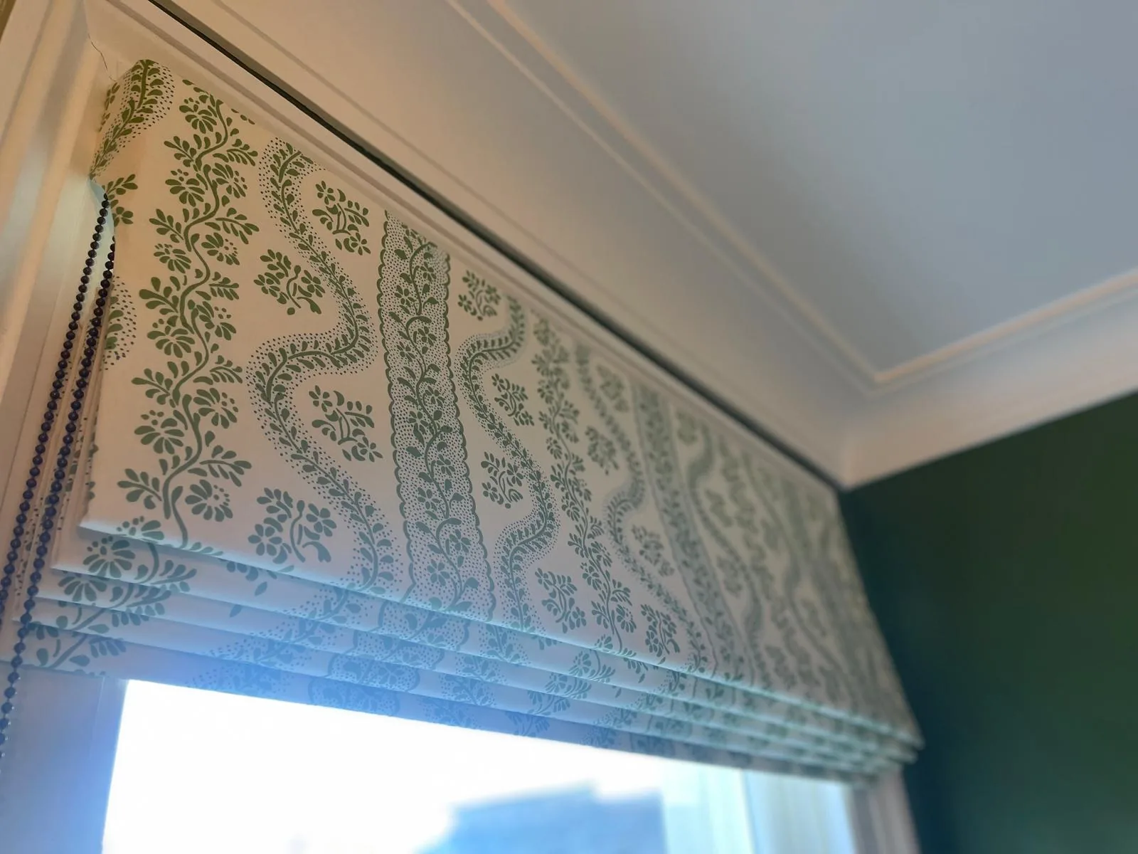 Green and white trellis-print Roman blind raised above a window on a dark green wall