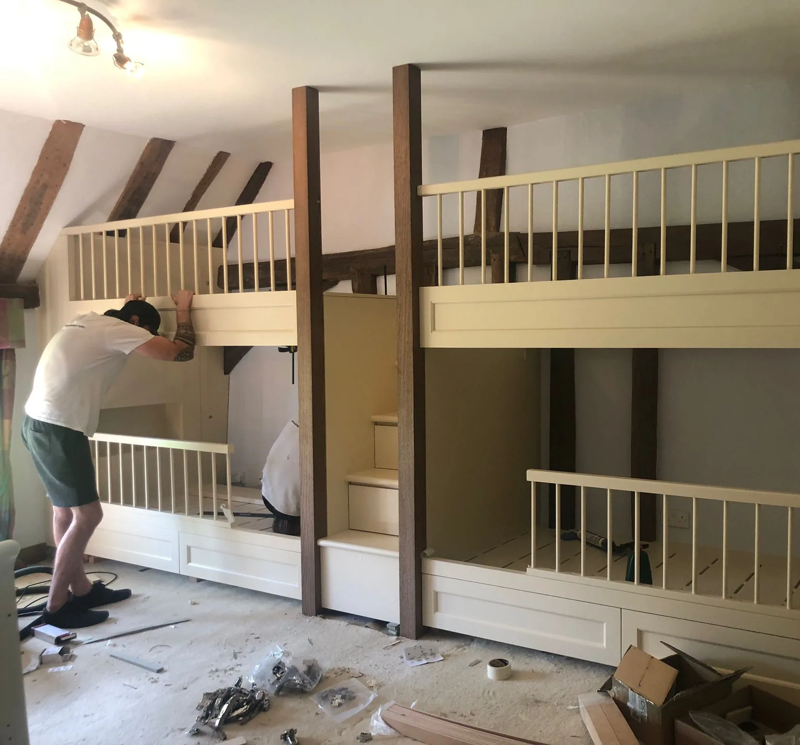 Painted bunk beds with timber posts and rails being installed under a beamed roof, with fitters at work and fixings on the floor