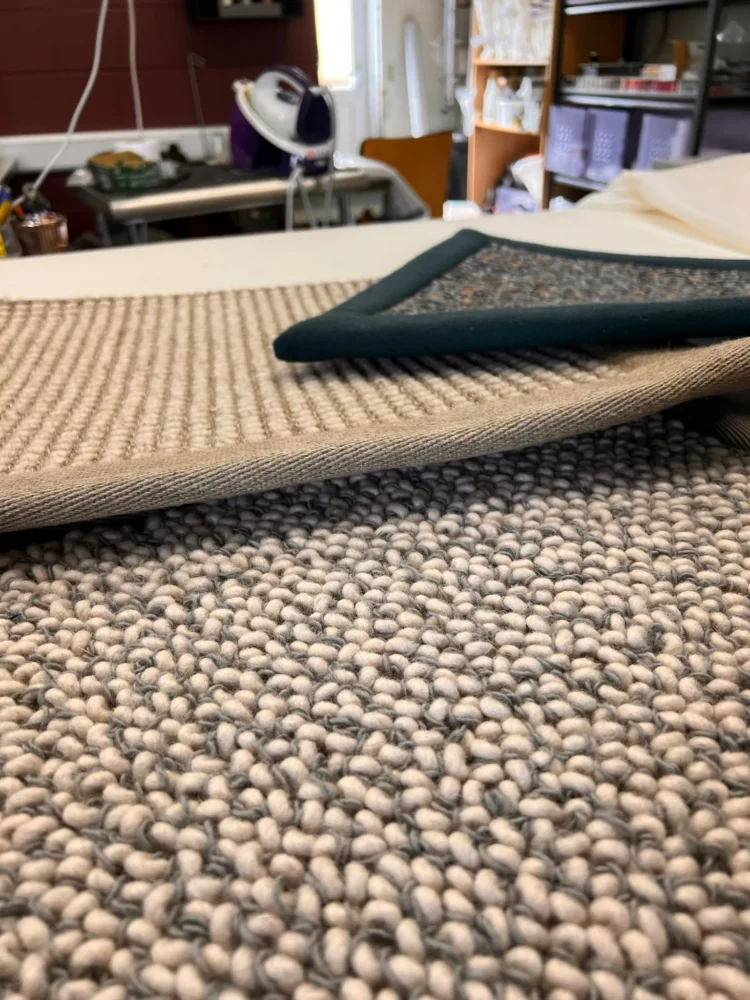 Close-up of a pebble-weave rug sample with a dark teal bound edge, on a workroom table