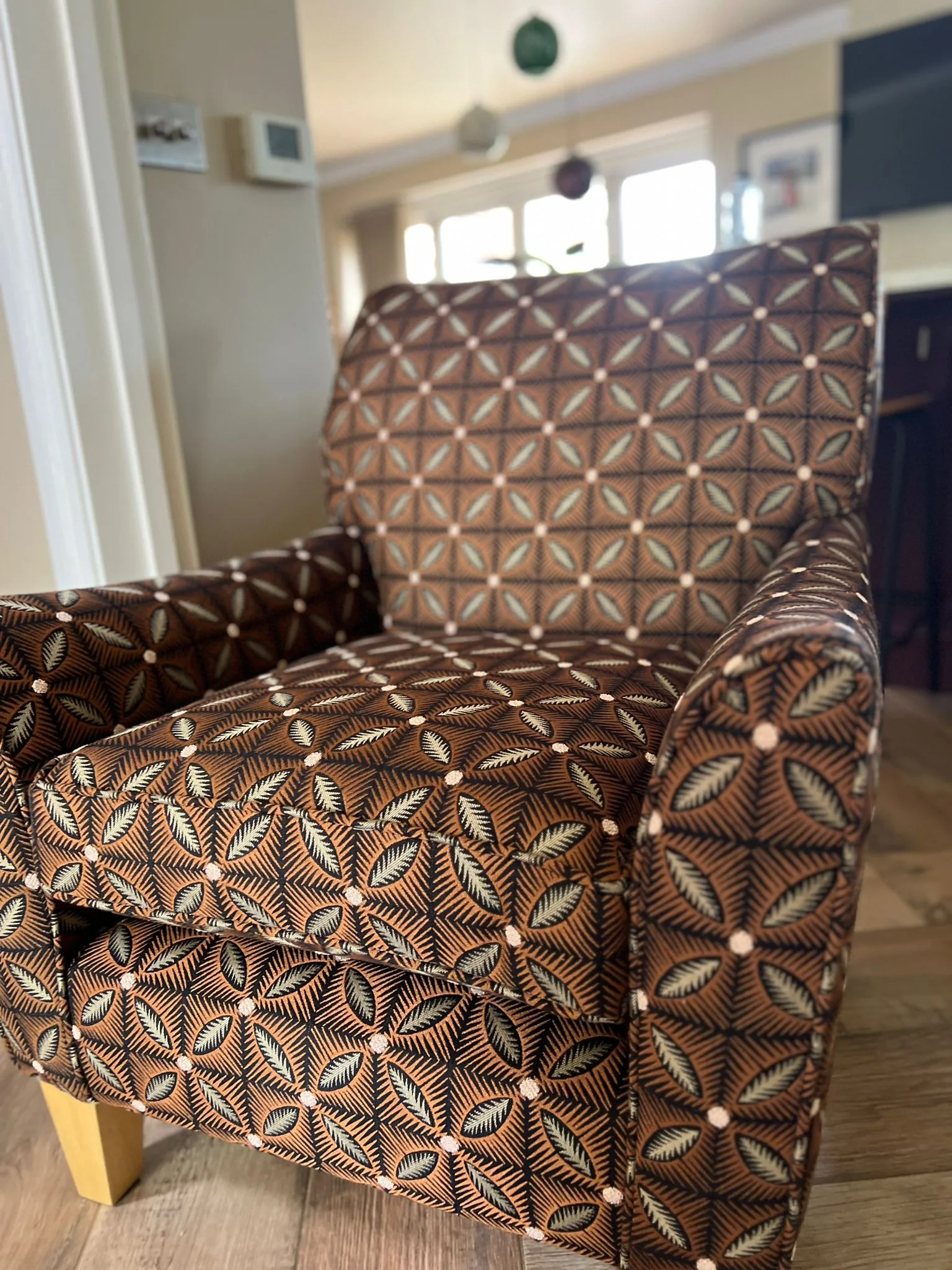 Armchair upholstered in a brown, teal and blush geometric leaf print, on pale wooden feet
