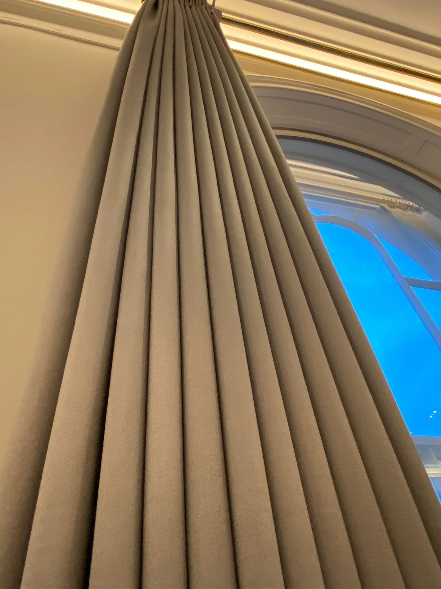 Looking up a tall taupe curtain beside an arched sash window, with cornice lighting above
