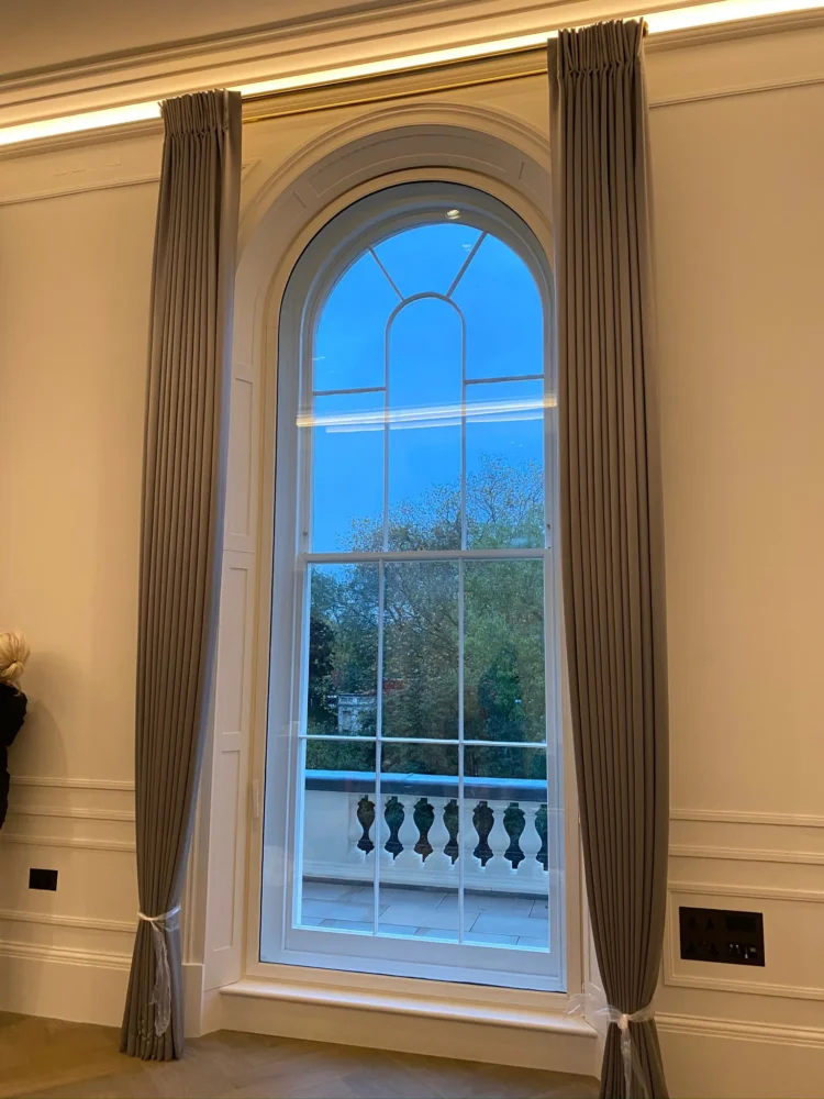 Tall taupe curtains with tiebacks framing an arched sash window that opens onto a balustraded terrace