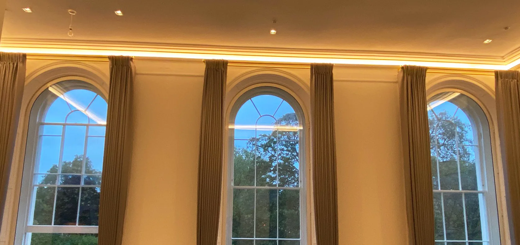 Three arched sash windows framed by tall taupe curtains, under a softly lit cornice